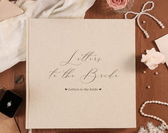May include: A beige linen-covered book titled "Letters to the Bride" with elegant script. The book is surrounded by wedding-themed accessories, including a ring box, pearls, and a rose.