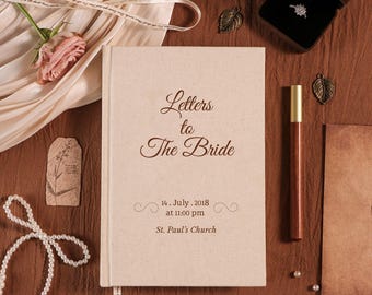May include: A beige linen-covered book titled "Letters to The Bride" with a gold pen, a ring in a box, and decorative elements. The book includes the date July 14, 2018, and the time and location of the wedding. Pearls and a rose are also visible.