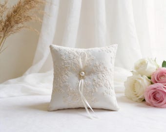 May include: Square white ring pillow with intricate ivory lace detailing. A faux pearl and ribbon accent the center. The pillow is set on a white surface, with white and pink roses in the background. A decorative item for a wedding.