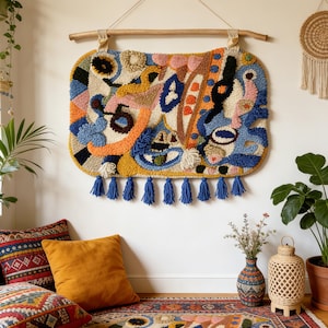 May include: A colorful, abstract wall hanging with a wooden rod. The textile features a mix of blue, orange, yellow, and pink shapes. Blue tassels hang from the bottom. Pillows and a rug with similar patterns are in the foreground.