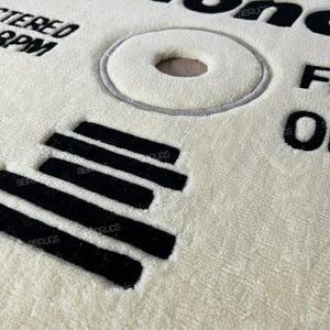 May include: Cream-colored round rug with black text and design elements. The rug features the word "blonde" in large letters, along with other text and a circular design resembling a vinyl record. Black parallel lines are also present.