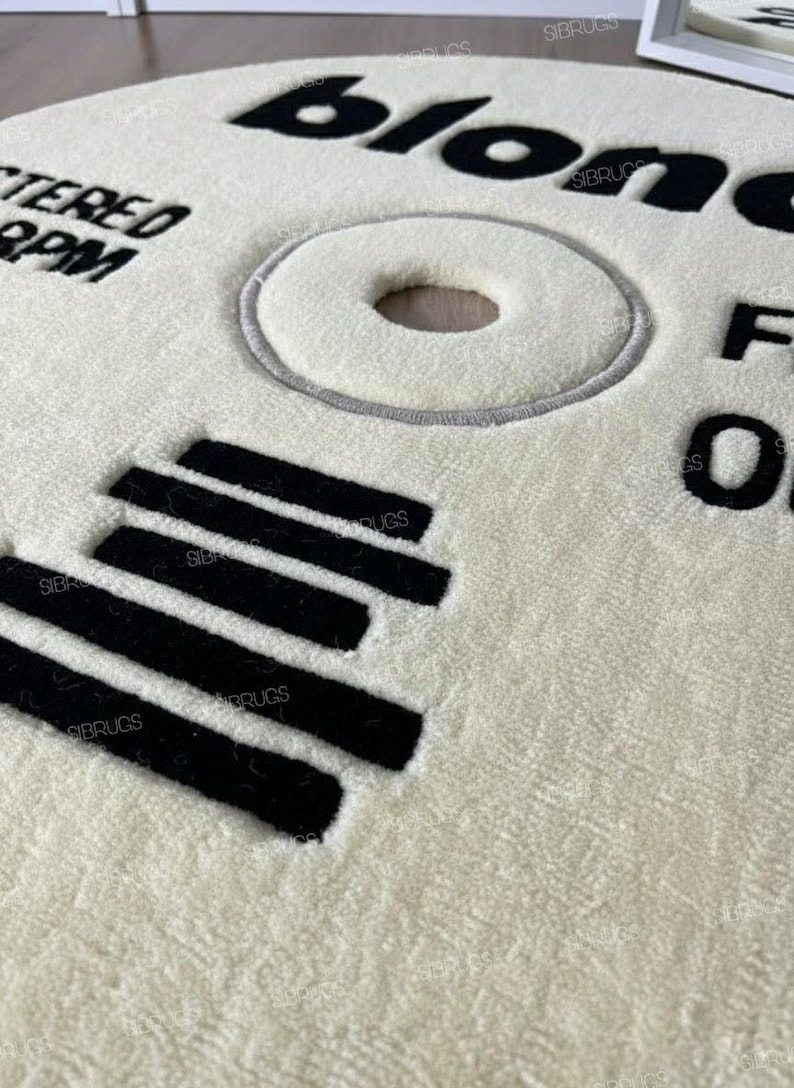 May include: Cream-colored round rug with black text and design elements. The rug features the word "blonde" in large letters, along with other text and a circular design resembling a vinyl record. Black parallel lines are also present.