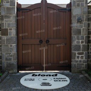 May include: A round, off-white rug with the text "blond FRANK OCEAN" in black, designed to resemble a vinyl record. The rug is placed in front of a dark wooden gate, flanked by stone pillars. The text "STEREO 33 RPM" is also visible.