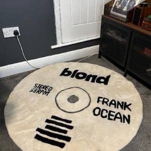 May include: Round, cream-colored rug designed to resemble a vinyl record. The rug features the word "blond" in black, along with "FRANK OCEAN" and "STEREO 33RPM". The design includes a central circle and black lines.