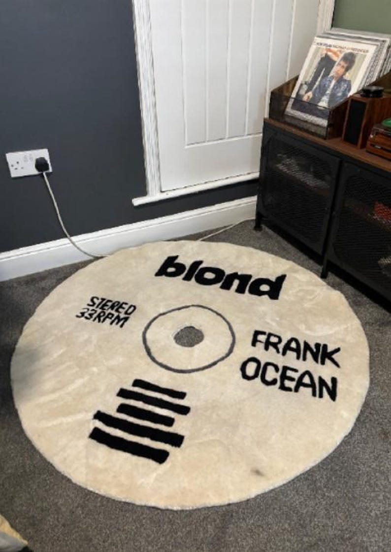 May include: Round, cream-colored rug designed to resemble a vinyl record. The rug features the word "blond" in black, along with "FRANK OCEAN" and "STEREO 33RPM". The design includes a central circle and black lines.