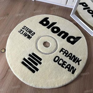 May include: A cream-colored, circular rug with black text that reads "blond", "STEREO 33 RPM", and "FRANK OCEAN". The rug is designed to resemble a vinyl record.