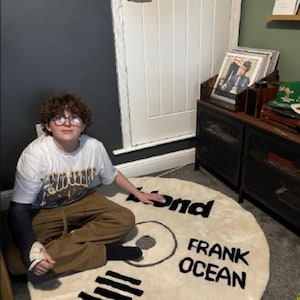 May include: A cream-colored, circular rug designed to resemble a vinyl record. The rug features the words "Blond" and "Frank Ocean" in black lettering. The rug is placed on a gray carpet.