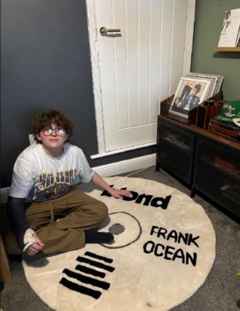 May include: A cream-colored, circular rug designed to resemble a vinyl record. The rug features the words "Blond" and "Frank Ocean" in black lettering. The rug is placed on a gray carpet.