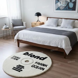 May include: A round, off-white rug with black text that reads "blond FRANK OCEAN STEREO 33 RPM". The rug is designed to resemble a vinyl record and is placed on a wooden floor in a bedroom setting.