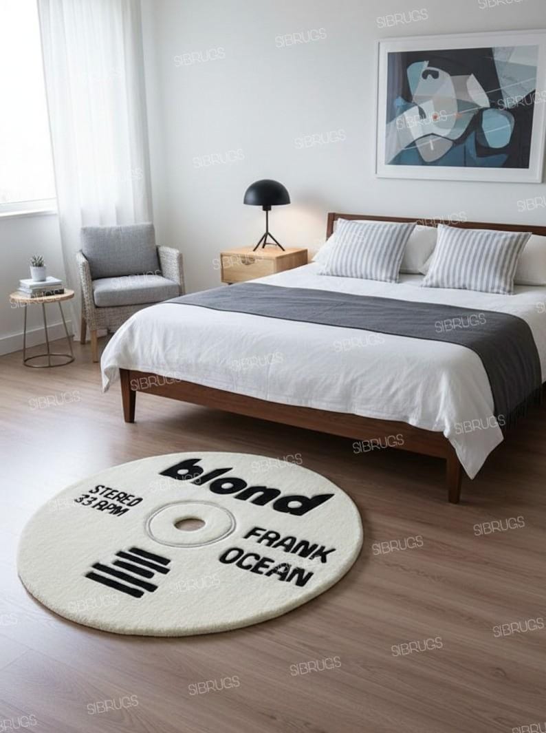 May include: A round, off-white rug with black text that reads "blond FRANK OCEAN STEREO 33 RPM". The rug is designed to resemble a vinyl record and is placed on a wooden floor in a bedroom setting.