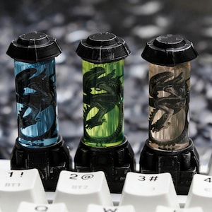 May include: Three custom keycaps with a black alien design inside cylindrical containers. The containers have blue, green, and amber hues, with black bases and tops. The keycaps are on a white keyboard.