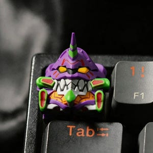 May include: A custom keycap featuring a purple, green, and orange robot design. The keycap is on a black keyboard, with the "Tab" and "F1" keys visible. The robot has a distinctive face and a horn.