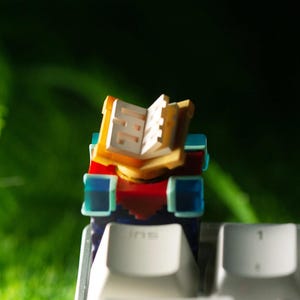 May include: A custom mechanical keyboard keycap featuring a pixelated open book design. The keycap has a red, blue, and yellow color scheme, with a white book interior. The keycap is set against a blurred green background.