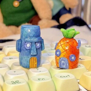 Op de afbeelding: Twee miniatuurbeeldjes: een blauw, steenachtig hoofd en een oranje ananas huis. De blauwe figuur heeft een rechthoofdige kop met cirkelvormige ogen en een kleine deur. Het ananashuis heeft een groene bovenkant en blauwe accenten. Beide staan op een toetsenbord.