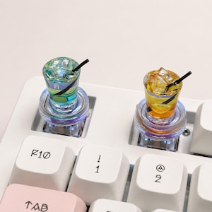May include: Two novelty keyboard keycaps shaped like miniature cocktails. One is blue and green, the other is yellow with a lemon slice. Both have black straws and sit on clear, illuminated bases. The keyboard keys are white.
