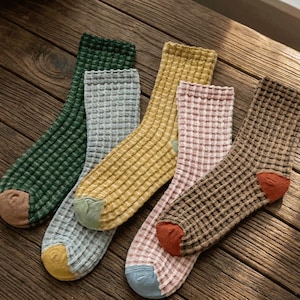 Pode incluir: Cinco pares de meias de tripulação texturizadas em várias cores, incluindo verde, cinza, amarelo, rosa e marrom. Cada meia tem um padrão de malha waffle e cores contrastantes nos dedos e calcanhar. As meias estão dispostas em uma superfície de madeira.