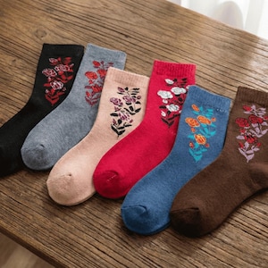 May include: Six pairs of crew socks in various colors, each adorned with floral embroidery. The socks are black, gray, pink, red, blue, and brown. The embroidered flowers feature shades of red, white, and orange.
