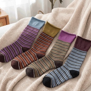 May include: Four pairs of striped socks in various colors: purple, brown, green, and blue. Each pair has a different colored cuff and toe. The socks are arranged on a beige blanket.