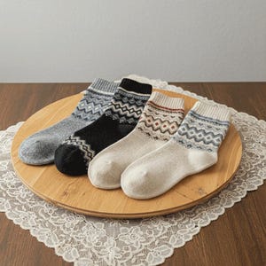 May include: Four pairs of patterned socks in grey, black, cream, and off-white, displayed on a wooden tray. Each sock features a geometric design in contrasting colours, with ribbed cuffs. The socks are made of a soft, warm material.
