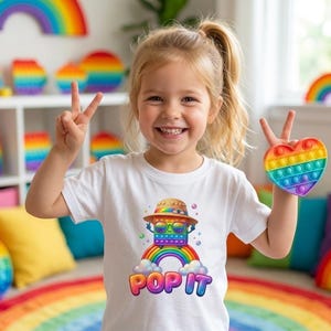 Puede incluir: Una niña sonriente sostiene un juguete pop-it en forma de corazón con los colores del arcoíris. Lleva una camiseta blanca con un gráfico colorido de un robot sobre un arcoíris, con las palabras "POP IT". El fondo presenta decoración de arcoíris.
