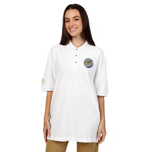 May include: White polo shirt with short sleeves and a collar. The shirt has a circular logo on the chest and a small design on the left sleeve. The logo is a blue and yellow design with the word "Bergen".