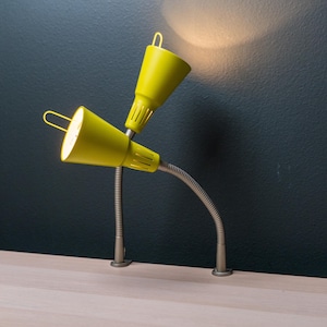 May include: A dual-head desk lamp featuring two bright yellow conical shades attached to flexible, metallic gooseneck arms. Each shade includes a small metal loop at the top and a vented base, with the lamp mounted on a flat surface.