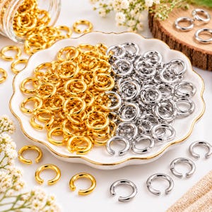May include: A collection of small, circular metal jump rings in gold and silver tones. The rings are arranged in a white scalloped dish, with some scattered around the base. Each ring features a small opening, indicating they are open jump rings.