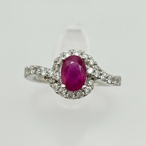 May include: A silver-toned ring featuring a central oval-cut deep pink ruby gemstone surrounded by a halo of small round white diamonds. The band has a bypass design that curves around the center stone, also set with small round white diamonds.