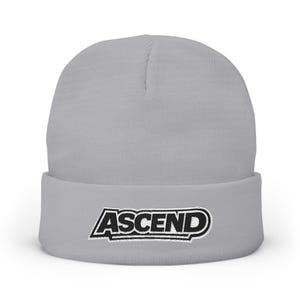 May include: A light grey beanie hat featuring the word "ASCEND" embroidered in black and white. The hat is made of a soft, knitted fabric and has a folded brim. The design is simple and modern, suitable for various styles.