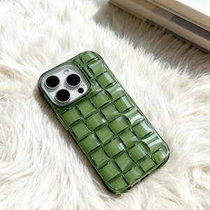 May include: A green phone case with a woven pattern. The case has a clear border around the camera lenses and a textured, grid-like design across the back. The case is shown on a white, fluffy surface.