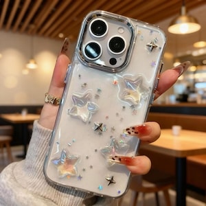 Op de afbeelding: Transparante telefoonhoes met een glitterend, sterren-thema ontwerp. De hoes heeft een transparante behuizing met stervormige versieringen in verschillende maten en een zilveren cameraring.