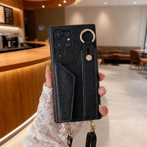 Puede incluir: Funda de teléfono de cuero negro con ranura para tarjetas y un anillo y correa de color dorado. La funda tiene una superficie texturizada y un botón dorado. Una correa negra está unida a la funda.