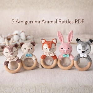 Peut inclure: Cinq hochets animaux amigurumi faits à la main, dont un cerf, un ours, un renard, un lapin et un loup. Chaque hochet est doté d'un anneau en bois et est crocheté dans des couleurs pastel douces avec des détails délicats. Le texte "5 Amigurumi Animal Rattles PDF" est visible.