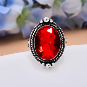 May include: A silver ring featuring a large, oval-cut, vibrant red gemstone set in a decorative frame. The setting includes a twisted rope-like border and small silver spheres at the top and bottom, creating a detailed, textured metal finish.