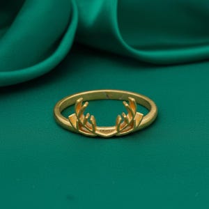 May include: A gold-colored ring with a deer antler design. The ring is set against a green background. The antlers are detailed and add a unique touch to the jewelry.