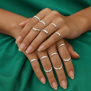 以下が含まれることがあります： さまざまなスタイルのシルバーリングのコレクション。カーブしたデザインやバンドデザインなどがあります。複数の指にリングを着用し、さまざまな形とサイズを表現しています。ジュエリーは緑色の背景に配置されています。