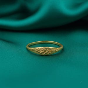 Puede incluir: Un anillo dorado con un diseño floral. El anillo tiene una banda lisa y redondeada y una cara ovalada ligeramente aplanada, adornada con un patrón en relieve de hojas y flores. El anillo está sobre un fondo turquesa.