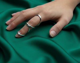 May include: A hand wearing two silver rings. One ring is a simple band, and the other is a multi-part ring that wraps around the finger. The background is a deep green fabric.
