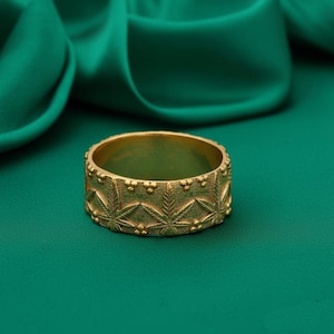 May include: A gold-colored ring with a repeating pattern of cannabis leaves. The ring is set against a green background, with a silky green fabric in the background. The ring is a band style, with a detailed design.