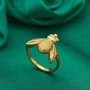 May include: A gold-toned ring featuring a three-dimensional bee ornament on the band. The bee has a textured, striped abdomen and smooth, extended wings that reach across the top of the ring. The metal has a polished, reflective finish.