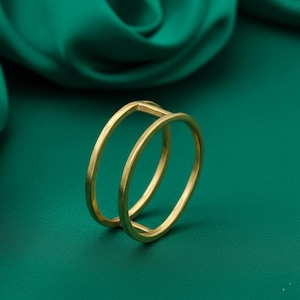 Puede incluir: Un anillo dorado con un diseño de doble banda. El anillo presenta dos bandas circulares paralelas conectadas por una pequeña barra recta. El anillo está sobre un fondo verde oscuro.