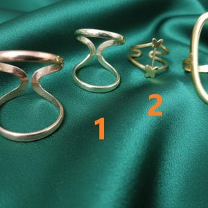 May include: Four rings of different designs are displayed on a green satin surface. The rings are made of gold-colored metal. One ring has a double-band design, another has a butterfly design, and a third has an oval shape.