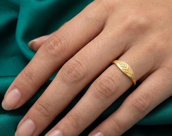 May include: A gold ring featuring a textured, matte finish and a delicate, engraved fern leaf pattern on the band. The ring has a tapered design that narrows towards the bottom, showcasing a detailed botanical motif across the top surface.