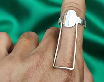May include: A silver ring with an oval top and a rectangular frame design. The ring is worn on a finger, showcasing its unique shape and metallic shine. The background is a solid green color.