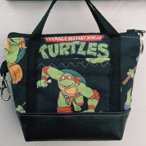 May include: A small, black tote bag featuring Teenage Mutant Ninja Turtles characters. The bag has a black base, a zipper closure, and a small pocket. The handles and trim are black, and the bag has a silver clasp.