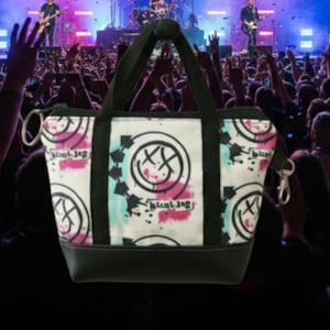 May include: A white tote bag with a black base and handles, featuring a repeating pattern of a smiley face with crossed-out eyes, arrows, and the text "blink-182" in black, pink, and teal. The bag has a zipper closure and a silver-colored metal ring and clasp.