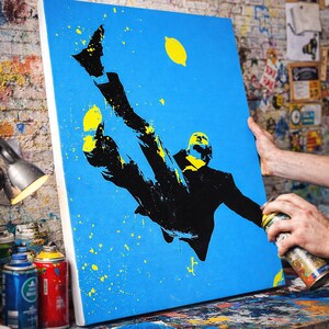 Puede incluir: Una obra de arte de plantilla hecha a mano sobre un lienzo azul. La obra representa una silueta negra de una persona con traje, en el aire, con acentos amarillos y salpicaduras de pintura. El texto "Handmade stencil artwork - each piece is unique" está en la parte inferior.