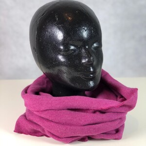 Könnte beinhalten: Ein magentafarbener Schlauchschal ist um einen schwarzen Schaufensterkopf drapiert. Der Schal besteht aus einem weichen, gestrickten Material und ist in mehreren Schichten gefaltet, wodurch seine Textur und Farbe zur Geltung kommen. Der Hintergrund ist eine schlichte weiße Oberfläche.