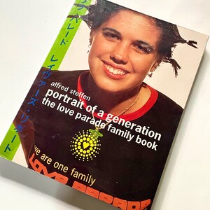 Puede incluir: Portada de libro con una persona que lleva una camiseta negra con el texto "portrait of a generation the love parade family book". También se ve texto japonés y las palabras "we are one family". La persona está sonriendo.