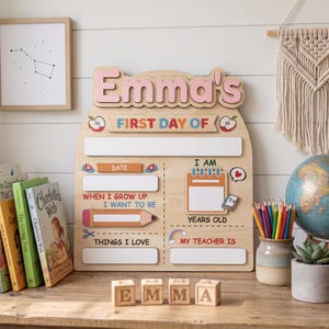 Könnte beinhalten: Ein Holzschild mit dem Text Emma's First Day Of, das leere Felder für Datum, Alter, Berufswünsche, Interessen und den Namen des Lehrers enthält. Das Brett zeigt bunte Grafiken von Äpfeln, einem Bleistift, einem Kalender und einem Regenbogen auf einer hellen Holzoberfläche.